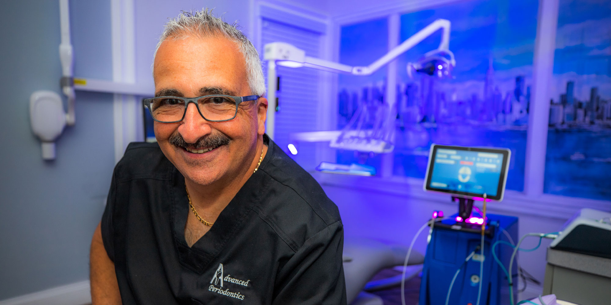 The image features a smiling man in a dental office setting. He has short gray hair, glasses, and a mustache. He is wearing a black dental uniform with "advanced Periodontics" embroidered on it. The background shows modern dental equipment and a screen displaying dental information, indicating a high-tech, professional dental practice. The ambiance of the room is enhanced by blue lighting, which adds a contemporary and clean feel to the environment. The man's friendly demeanor and the advanced equipment suggest a welcoming and well-equipped dental clinic.